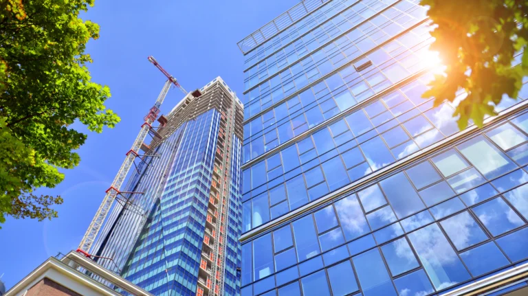 Glass office towers and a construction crane under a blue sky, with sunlight reflecting off the glass