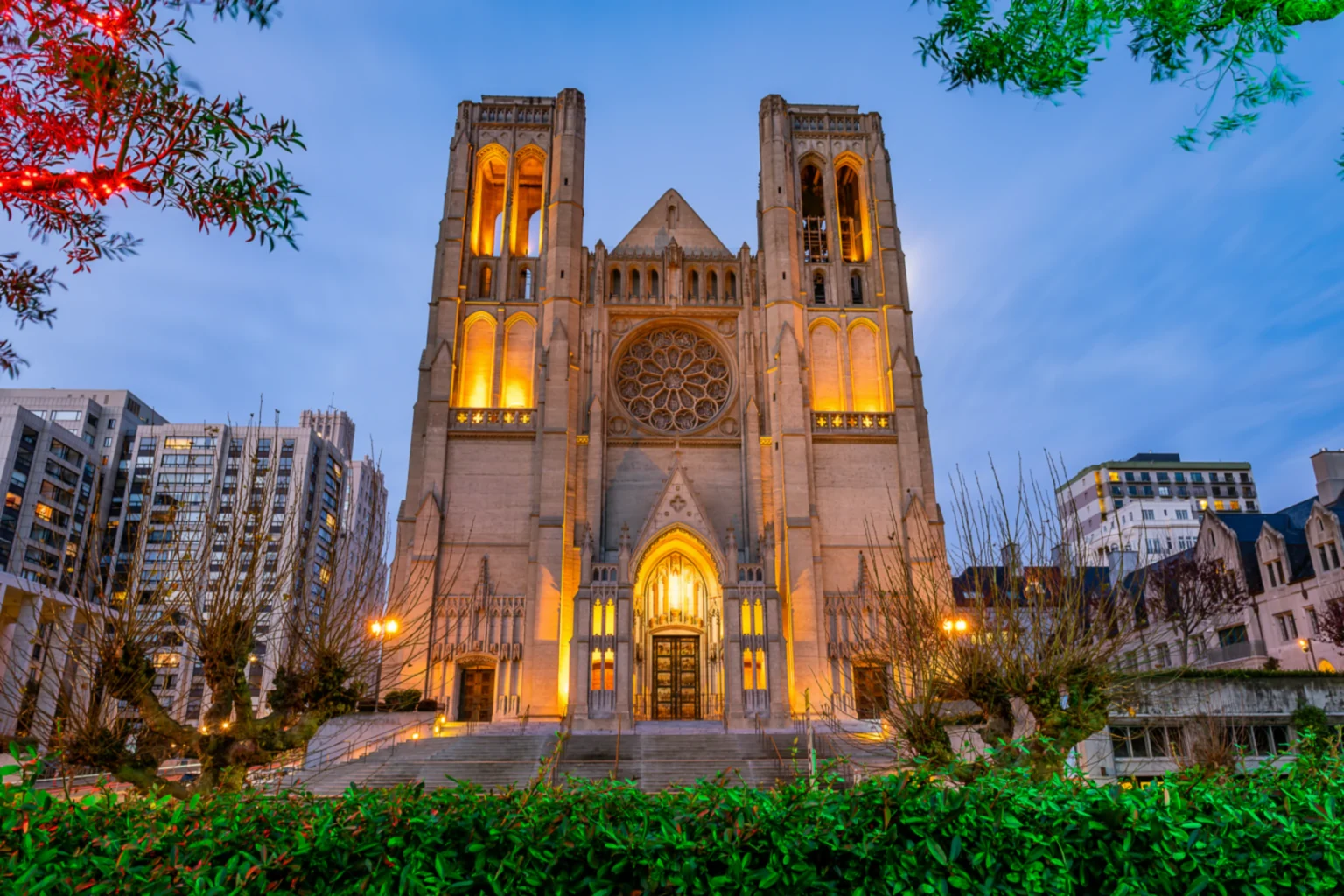 A Gothic cathedral facade lit at dusk, with a rose window and twin towers between city buildings