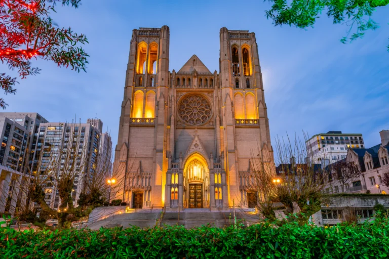 A Gothic cathedral facade lit at dusk, with a rose window and twin towers between city buildings