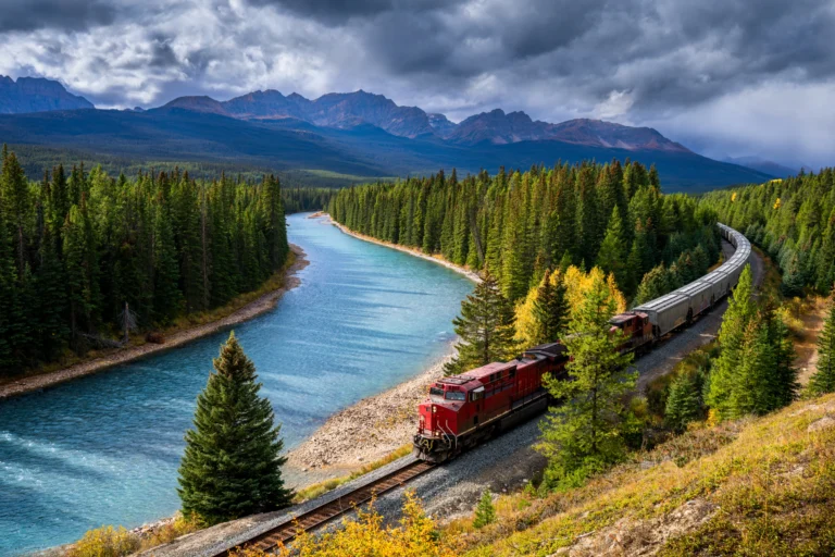 A red freight train curving along a turquoise river through evergreen forest, with mountains under storm clouds